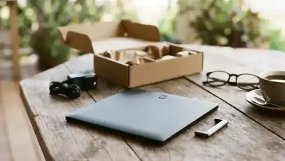 The back of a digital photo frame with a magnetic stand, a small power adapter and a coiled cord laid out on a wooden porch table beside a cardboard box